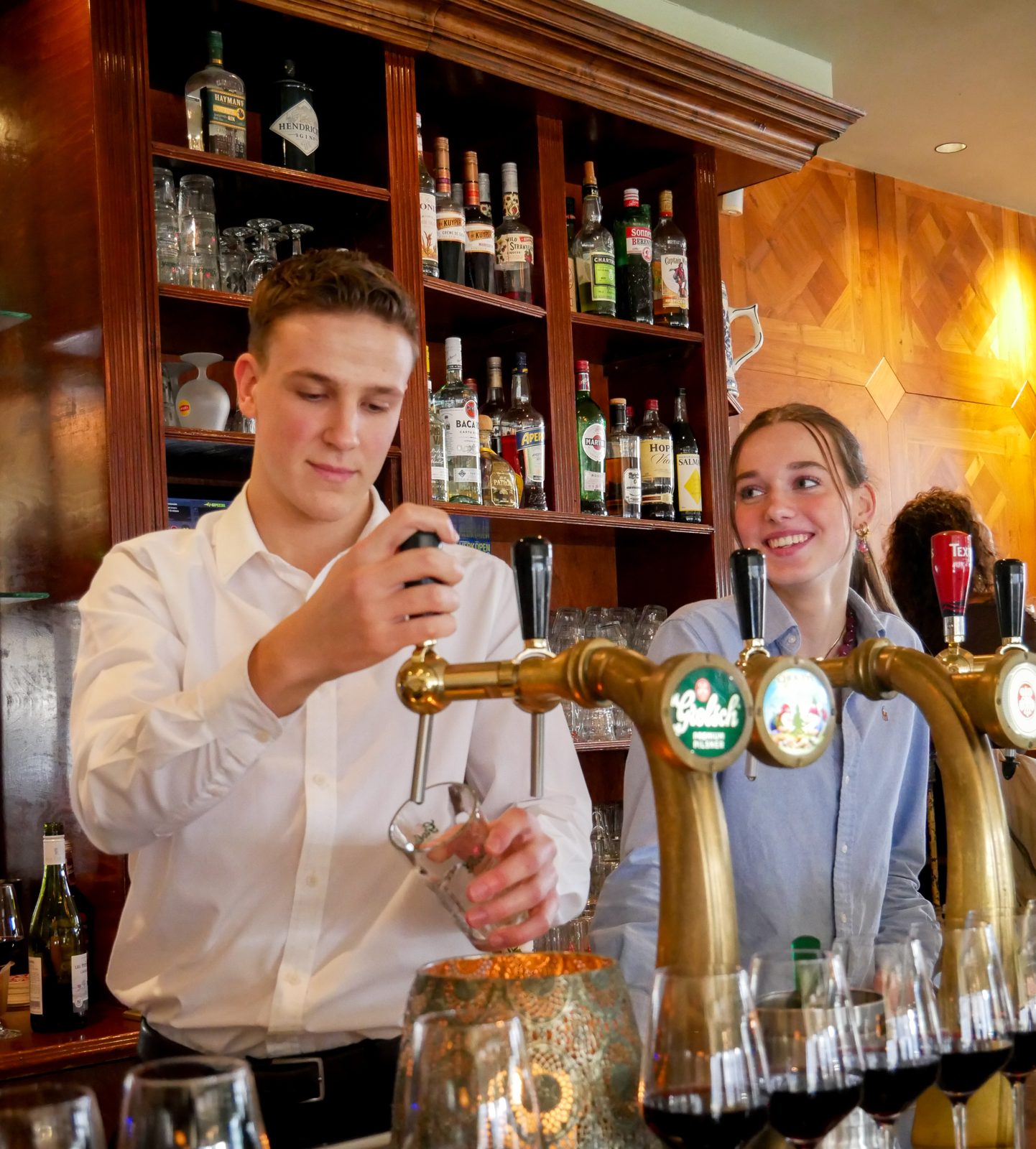 Twee medewerkers tappen bier achter de bar van De Vriend, met de flessenkast erachter