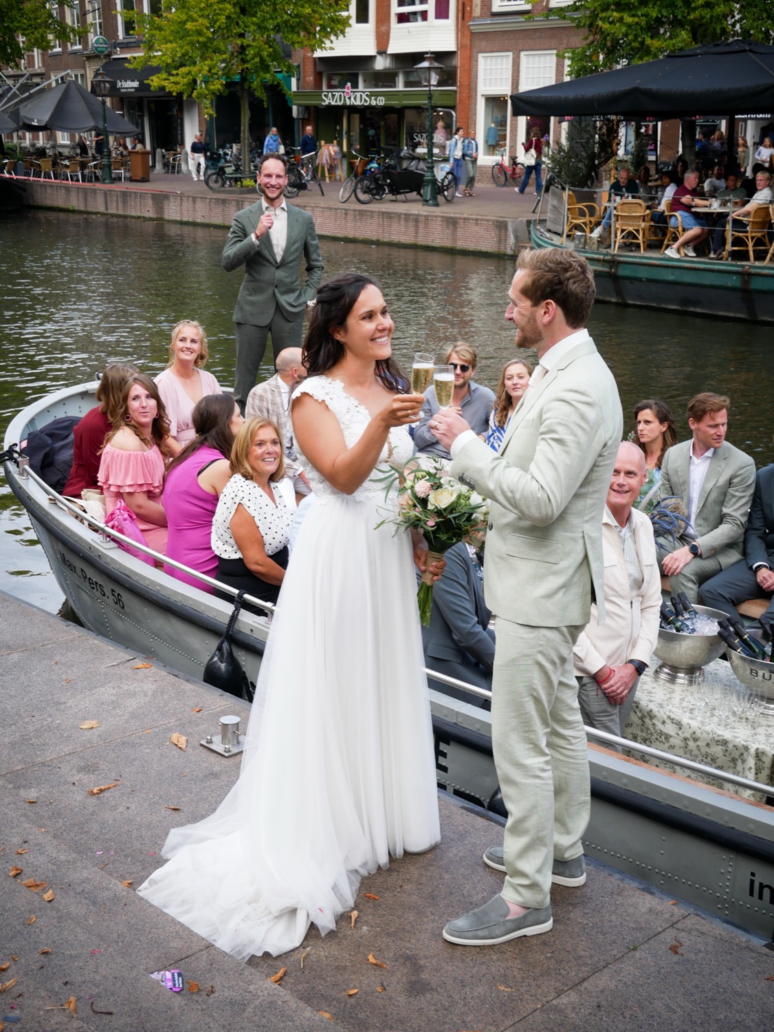Een bruidspaar proost met champagne op de kade, met de sloep vol gasten en de Leidse gracht op de achtergrond