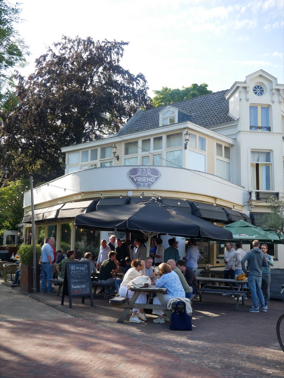 Het hoekpand van De Vriend met het uithangbord boven het terras, in zacht avondlicht