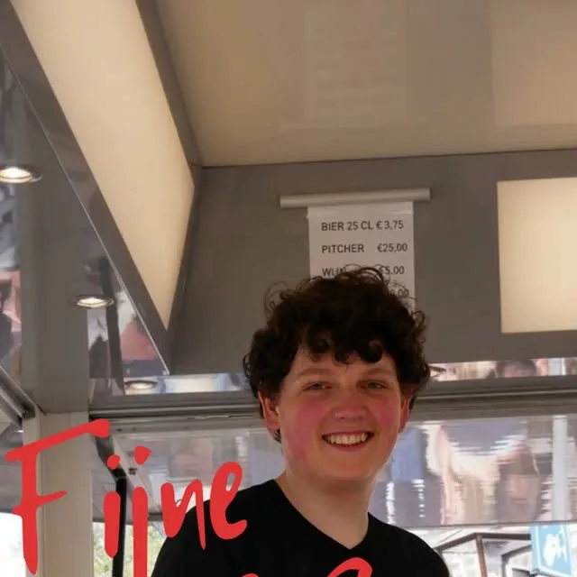 Een medewerker van De Vriend lacht achter de bar, met de prijzen voor bier op het bord erachter