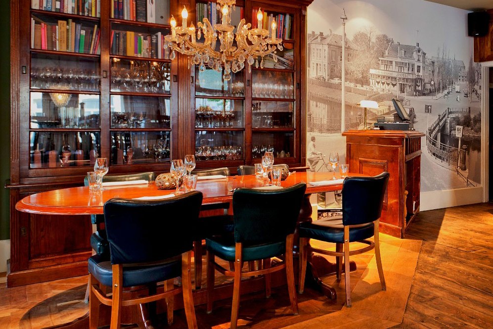 Het interieur van De Vriend: de gedekte tafel bij de boekenkasten, onder de kroonluchter, met de oude foto van Leiden op de muur