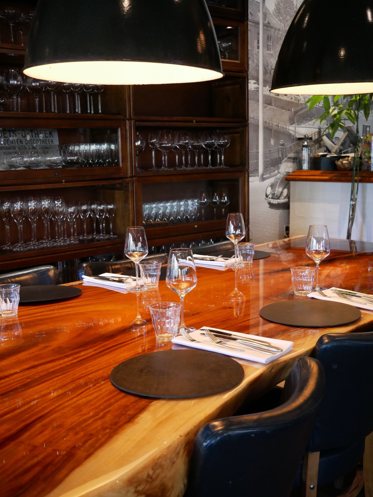 Het interieur van De Vriend: een lange houten tafel met gedekte plaatsen onder grote lampen, met de glazenkast op de achtergrond