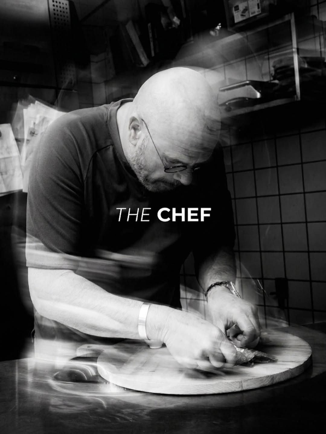 Jaap de Vriend, gebogen over een snijplank in de keuken, zwart-witfoto