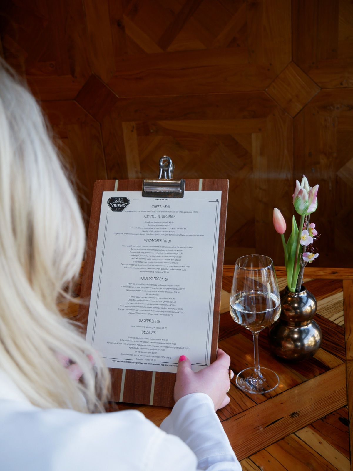 Een gaste leest de menukaart van De Vriend aan een houten tafel, met een glas witte wijn en tulpen
