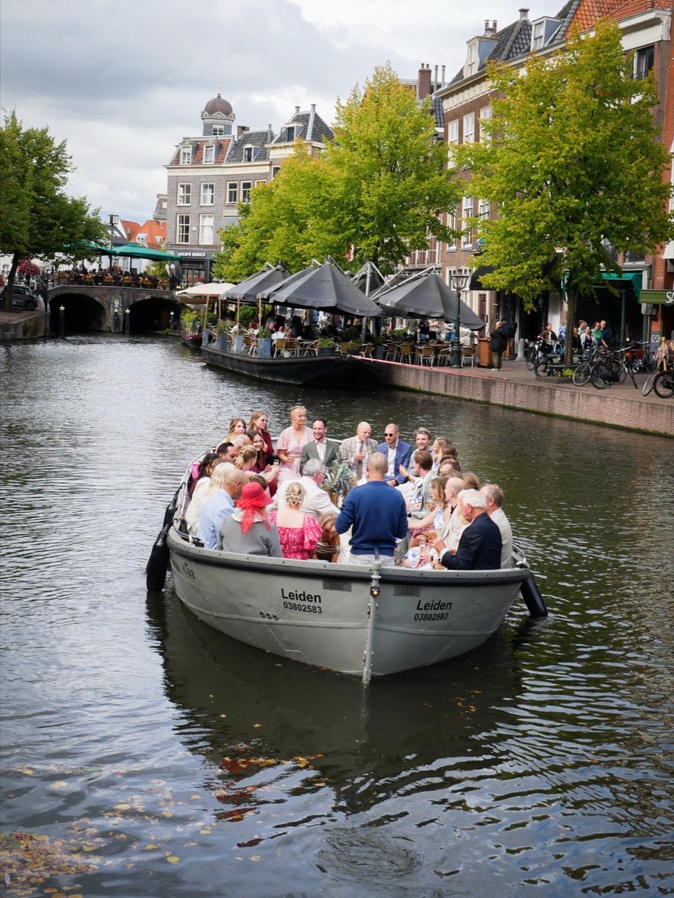 Een volle sloep met een gezelschap vaart over de Leidse gracht, met terrassen en gevels op de achtergrond