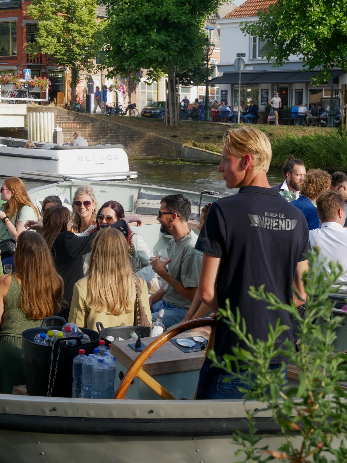 De schipper van Sloep de Vriend aan het roer, met het gezelschap en de kade op de achtergrond