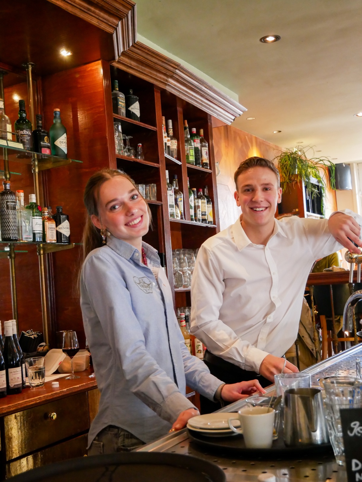 Twee medewerkers achter de bar van De Vriend, met de drankkast en het warme hout erachter