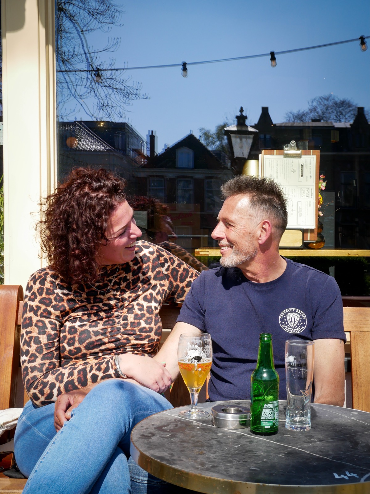 Een stel lacht naar elkaar aan een tafeltje op het terras van De Vriend, met speciaalbier op tafel