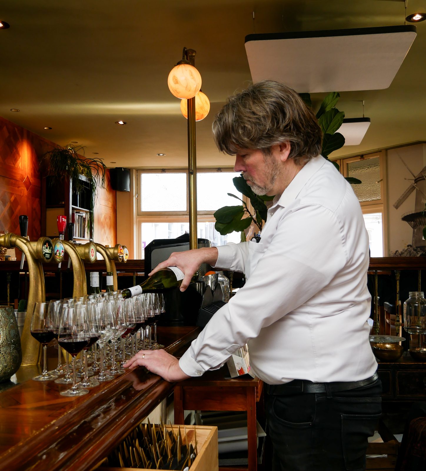 Een medewerker schenkt wijn in aan de bar van De Vriend, met een rij glazen klaar voor een gezelschap
